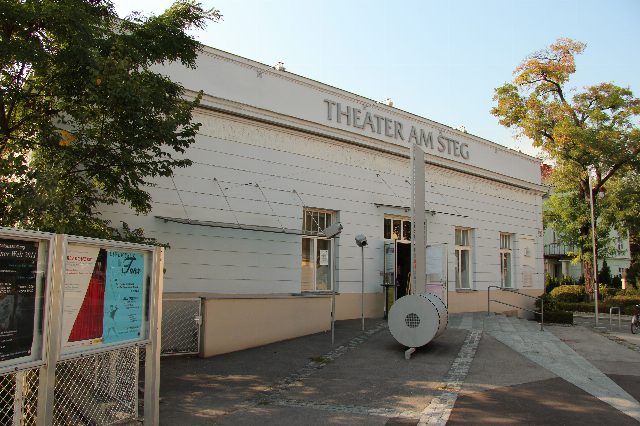 Weißes, langgestrecktes Gebäude mit der Aufschrift Theater am Steg, davor ein Vorplatz mit Plakatvitrinen und einer runden weißen Skulptur