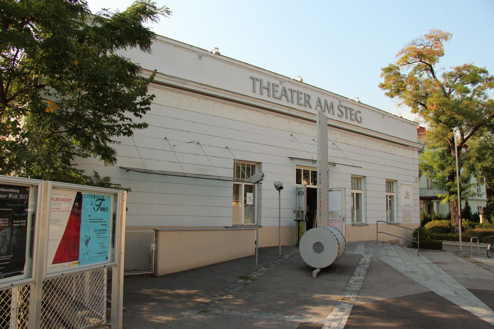 Weißes, langgestrecktes Gebäude mit der Aufschrift Theater am Steg, davor ein Vorplatz mit Plakatvitrinen und einer runden weißen Skulptur