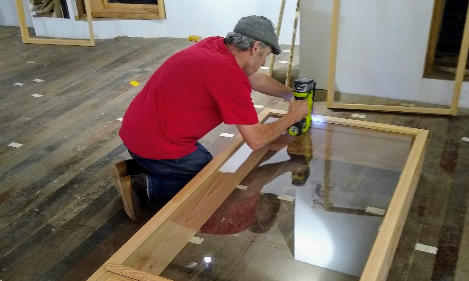 Handwerker in rotem T-Shirt kniet auf einem Holzboden und arbeitet mit einem Akkuschrauber an einem großen Holzfensterrahmen mit Glas
