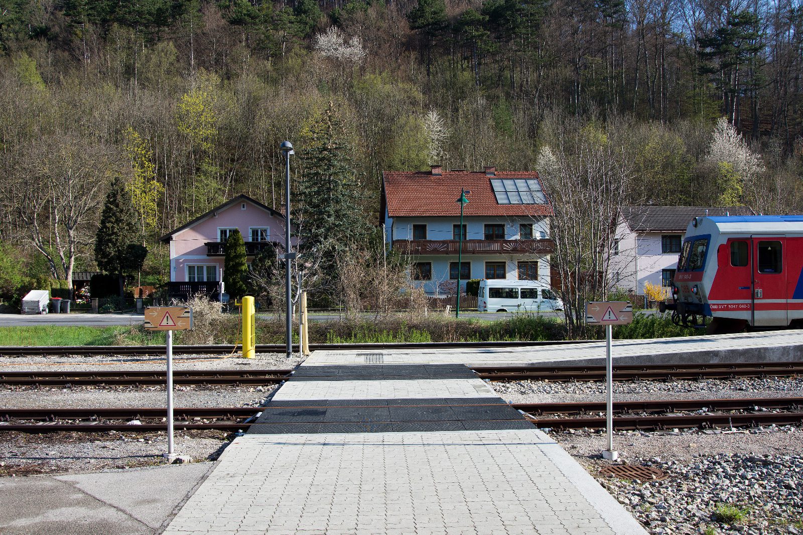 Bahnübergang am Bahnhof Weissenbach-Neuhaus, rechts ein rot-weißer Triebwagen der ÖBB, dahinter Wohnhäuser und bewaldeter Hang
