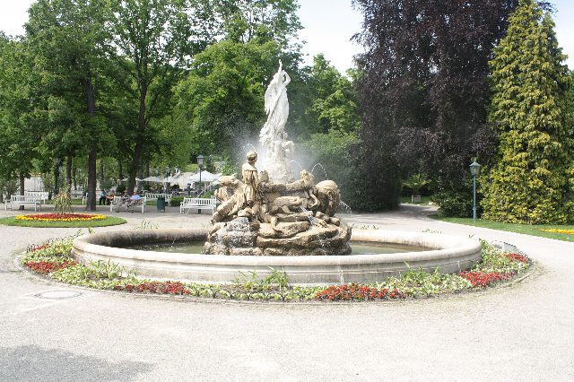 Undinebrunnen im Kurpark Baden mit Wasserfontänen, Blumenbeet und hohen Laubbäumen dahinter