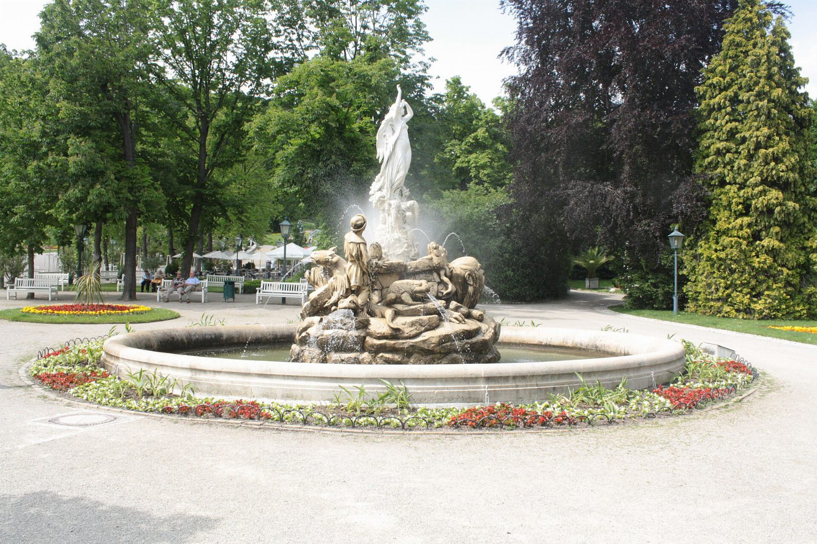 Undinebrunnen im Kurpark Baden mit Wasserfontänen, Blumenbeet und hohen Laubbäumen dahinter