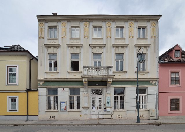 Dreistöckiges helles Gründerzeithaus mit goldenen Ornamenten und Balkon über dem Eingang, links ein gelbes, rechts ein rosa Nachbarhaus