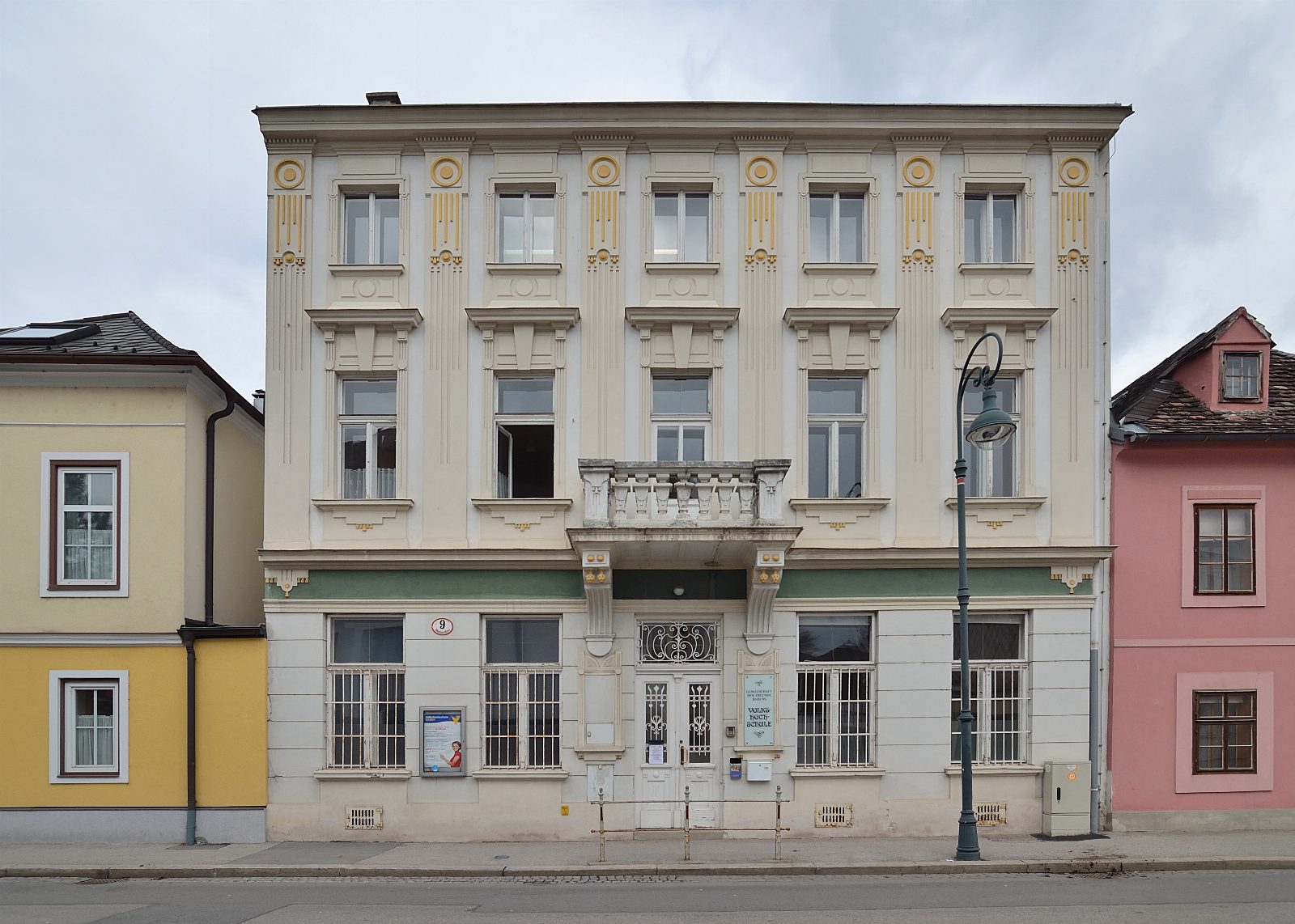 Dreistöckiges helles Gründerzeithaus mit goldenen Ornamenten und Balkon über dem Eingang, links ein gelbes, rechts ein rosa Nachbarhaus