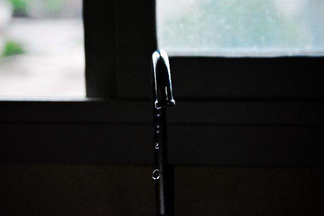 Tropfender Wasserhahn aus Chrom vor einem Fenster, darunter fallen einzelne Tropfen