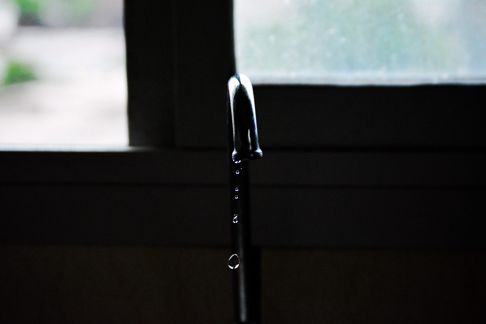 Tropfender Wasserhahn aus Chrom vor einem Fenster, darunter fallen einzelne Tropfen