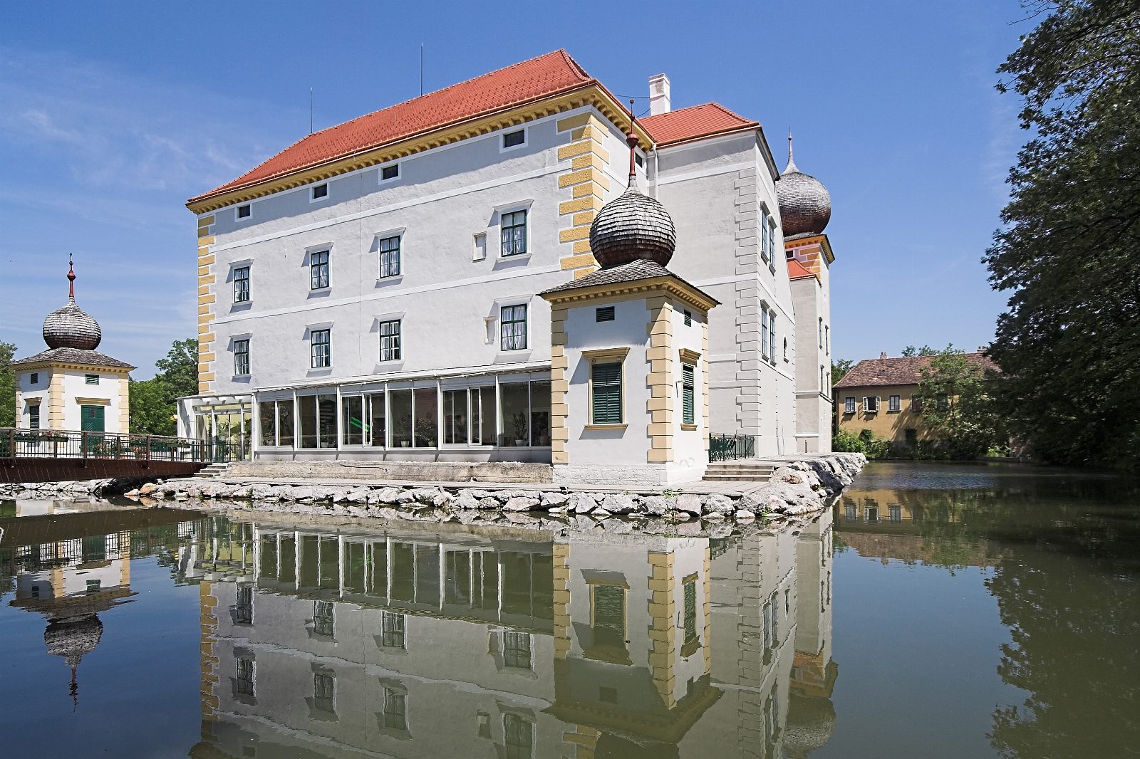 Wasserschloss Kottingbrunn mit weißer Fassade, rotem Dach und Zwiebeltürmchen, das sich im Schlossteich spiegelt