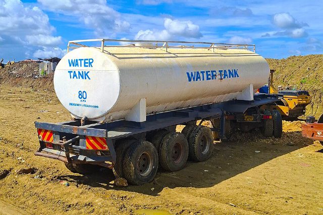 Ein weißer Tankanhänger mit der blauen Aufschrift Water Tank steht auf lehmigem Boden, dahinter ein Erdwall und blauer Himmel mit Wolken