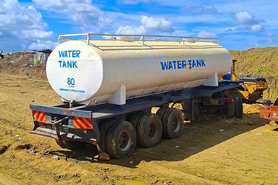 Ein weißer Tankanhänger mit der blauen Aufschrift Water Tank steht auf lehmigem Boden, dahinter ein Erdwall und blauer Himmel mit Wolken