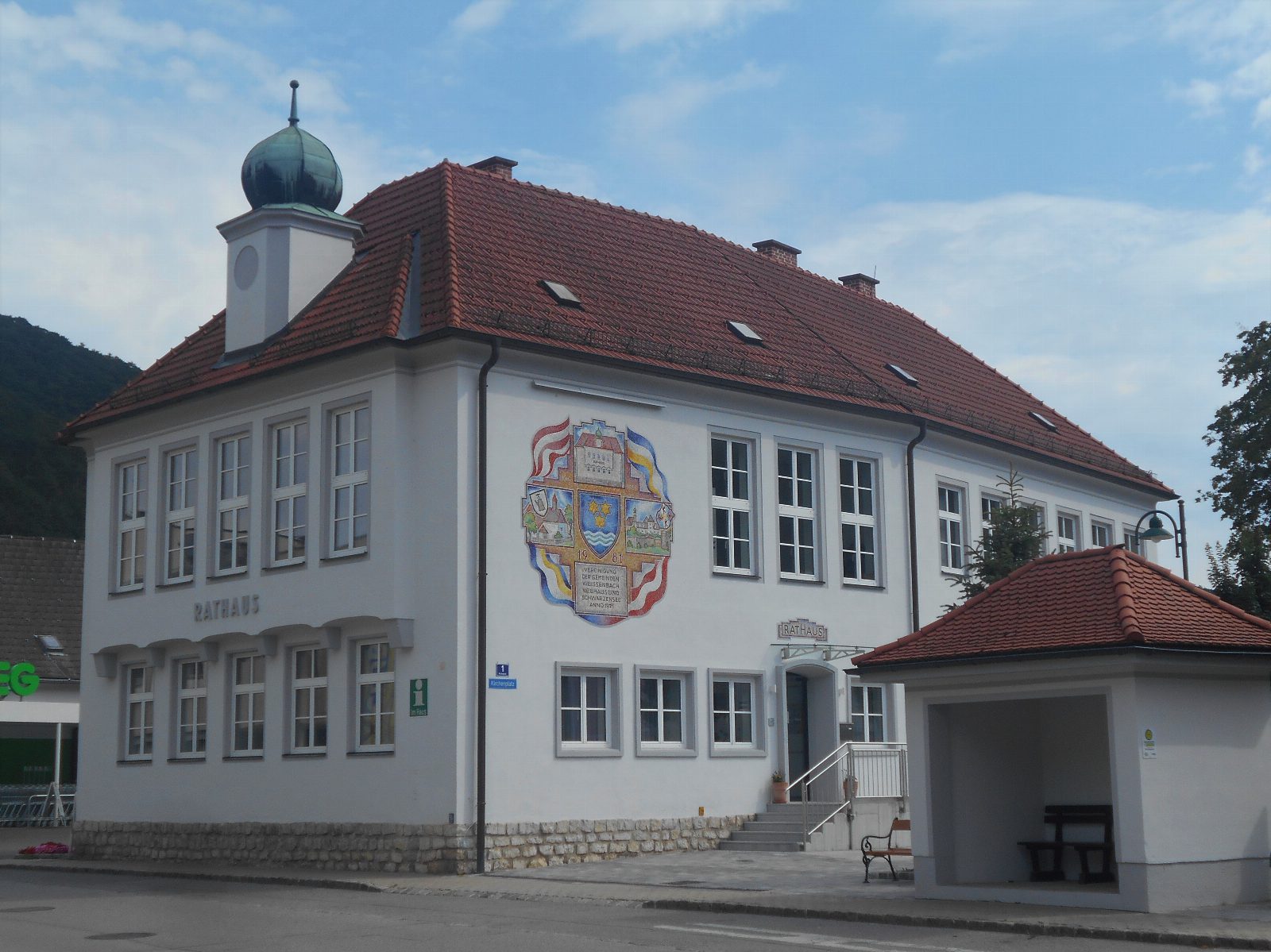 Weißes zweistöckiges Rathaus mit rotem Ziegeldach, Zwiebelturm und großem Wappen an der Fassade, rechts ein Buswartehäuschen