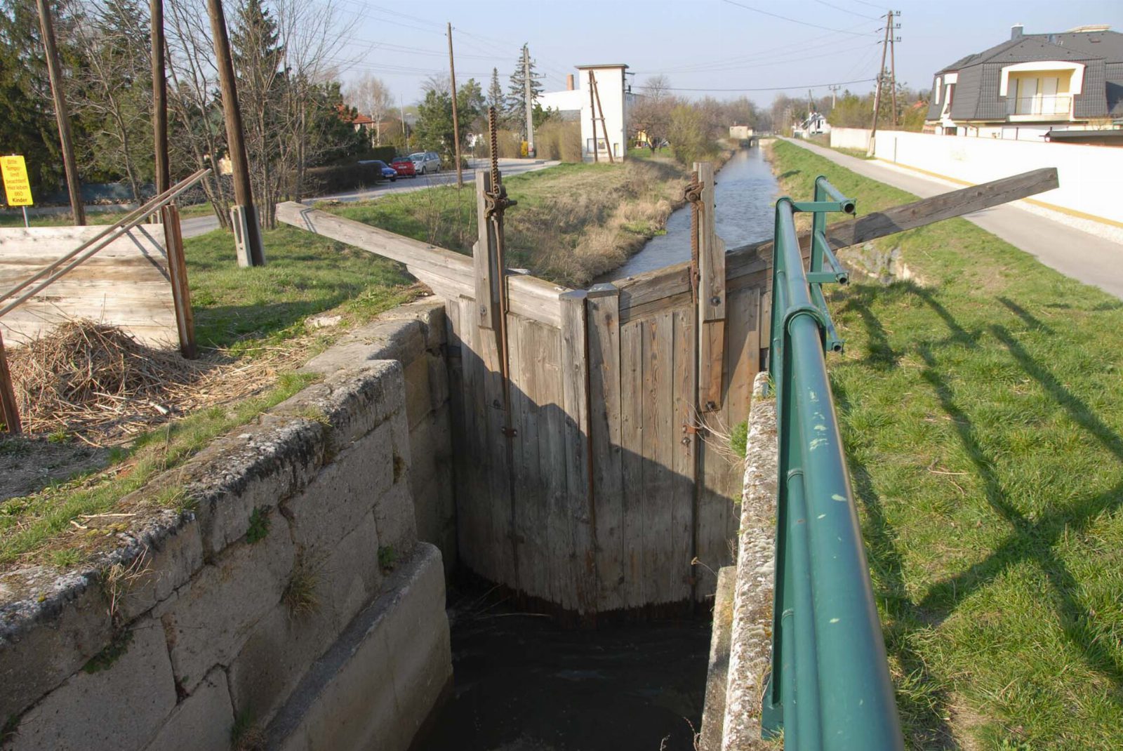 Hölzernes Schleusentor am Wiener Neustädter Kanal, daneben ein Geh- und Radweg und Häuser