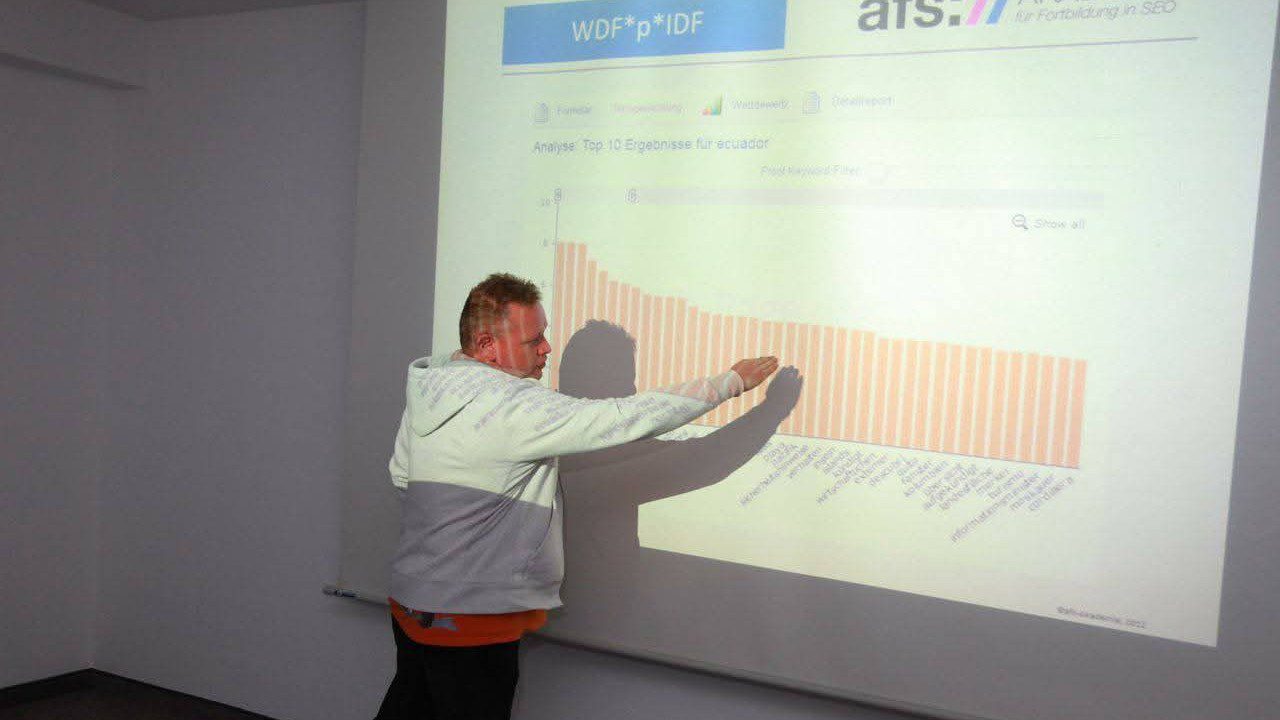 Ein Vortragender steht vor einer Projektionswand und zeigt auf ein Balkendiagramm einer Suchbegriff-Analyse