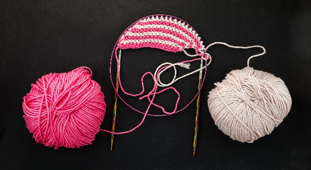 Ein rosa und ein weißes Wollknäuel auf schwarzem Grund, dazwischen ein gestreiftes Strickstück auf einer Rundstricknadel