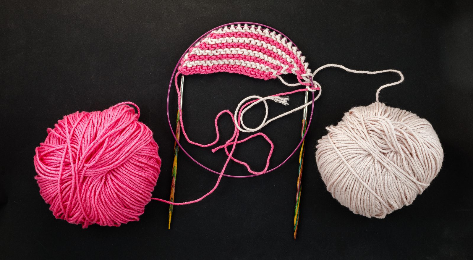 Ein rosa und ein weißes Wollknäuel auf schwarzem Grund, dazwischen ein gestreiftes Strickstück auf einer Rundstricknadel