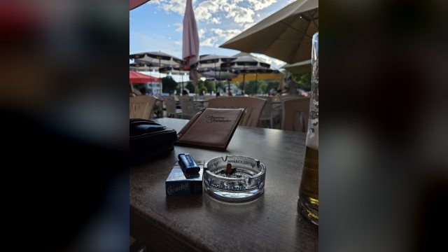 Zigarettenpackung mit Feuerzeug neben einem gläsernen Aschenbecher auf einem Tisch im Gastgarten, dahinter Sonnenschirme, rechts ein Bierglas