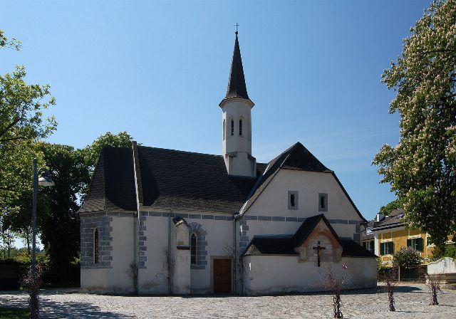 Bürgerspitalskirche in Enzesfeld: weiße Kirche mit spitzem, dunklem Turmhelm und Kruzifix am Anbau, davor ein gepflasterter Platz