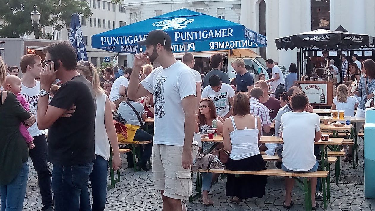 Besucherinnen und Besucher an Biertischgarnituren bei einem Fest im Freien, dahinter Getränke- und Imbissstände