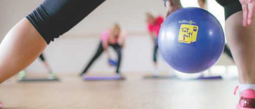 Hand auf einem lila Gymnastikball mit Tut-gut-Aufkleber, im Hintergrund trainieren Frauen auf Matten
