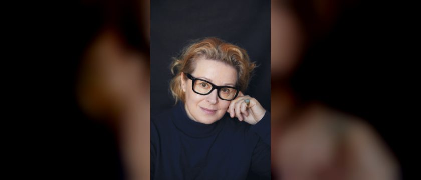 Porträt von Petra Morzé mit rotblonden Haaren, schwarzer Brille und dunklem Rollkragenpullover, die Hand an der Wange