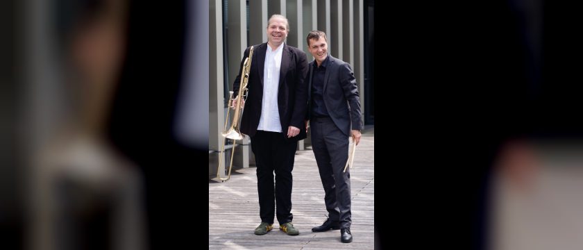 Markus Geiselhart mit Posaune und David Panzl mit Schlagzeugsticks stehen lachend auf einem Holzsteg