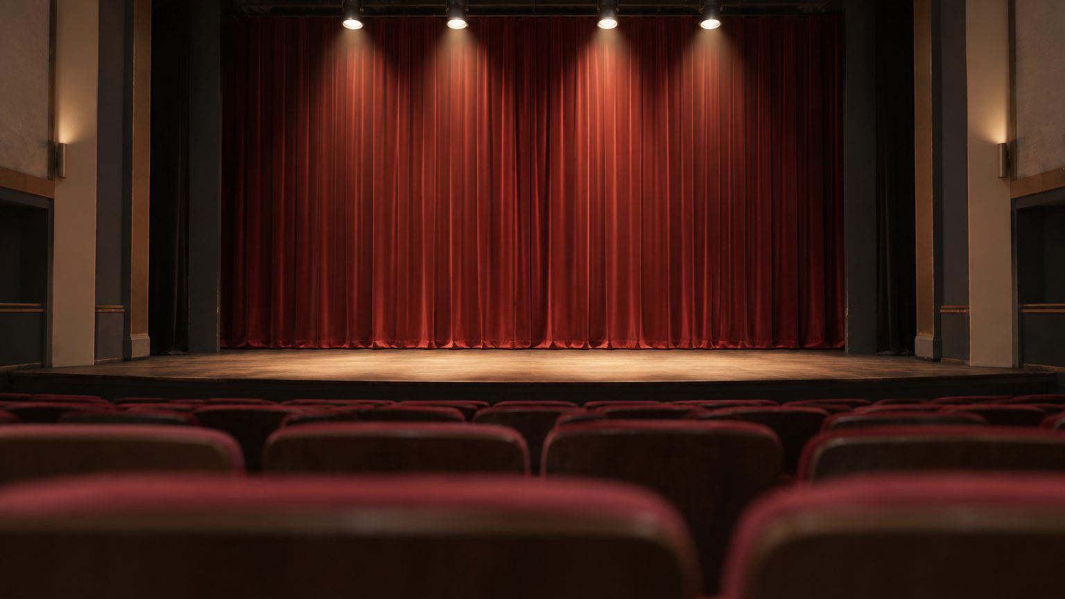 Symbolbild: der leere Zuschauerraum eines Stadttheaters, Blick über rote Sitzreihen auf einen beleuchteten roten Bühnenvorhang