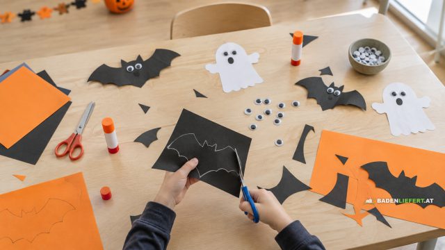 Symbolbild: Kinderhände schneiden Fledermäuse aus schwarzem Papier, auf dem Basteltisch Scheren, Kleber und Wackelaugen
