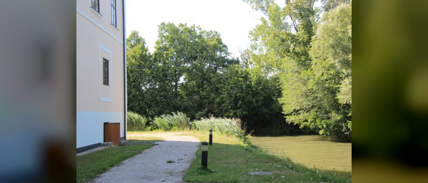 Kiesweg an der hellen Fassade von Schloss Vösendorf, daneben Wiese, Wasser und dichte Bäume im Sommer