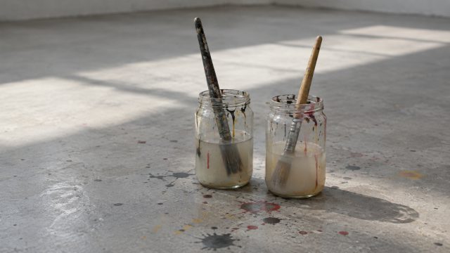 Symbolbild: zwei Gläser mit trübem Pinselwasser und Borstenpinseln auf einem Betonboden, daneben getrocknete Farbspritzer